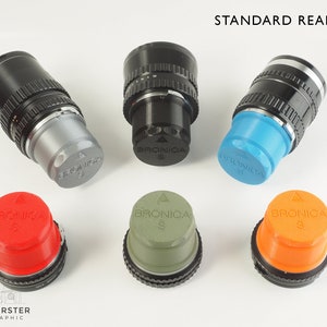 Op de afbeelding: Zes verschillende gekleurde achterdeksels voor een Bronica S-camera lens. De deksels zijn rood, blauw, groen, oranje, grijs en zwart. De deksels zijn allemaal van plastic en hebben het Bronica S-logo erop.