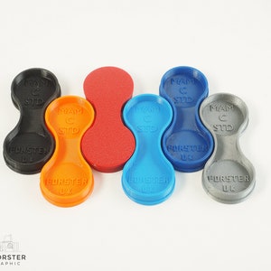 May include: Six 3D printed camera lens caps in different colours: black, orange, red, blue, grey, and a lighter grey. Each cap has the text "MAM C STD" and "FORSTER UK" printed on it.