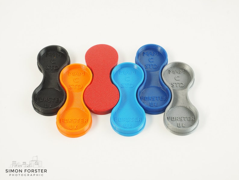May include: Six 3D printed camera lens caps in different colours: black, orange, red, blue, grey, and a lighter grey. Each cap has the text "MAM C STD" and "FORSTER UK" printed on it.