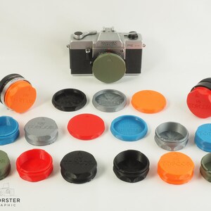 Può includere: Una collezione di 14 tappi per obiettivi di diversi colori per una fotocamera. I tappi per obiettivi sono tutti rotondi e hanno la scritta "Topcor" stampata su di essi. I tappi per obiettivi sono disposti in un cerchio attorno a una fotocamera argentata.