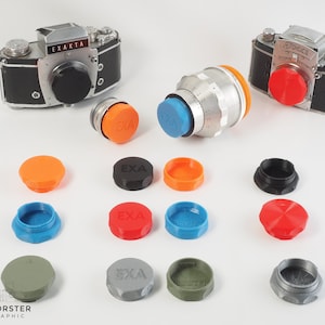 Può includere: Una collezione di tappi per obiettivi di fotocamera in vari colori, tra cui nero, grigio, blu, arancione e rosso. Alcuni tappi hanno la scritta "Exakta" stampata su di essi. I tappi sono disposti su una superficie bianca.