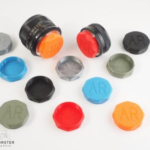 May include: A collection of 10 colorful lens caps with the letters "AR" printed on them. The caps are made of plastic and come in a variety of colors, including red, orange, blue, green, and gray. The caps are designed to fit over the lens of a camera.