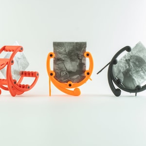 Op de afbeelding: Drie fotostandaards, één oranje, één zwart en één oranje, die elk een zwart-witfoto vasthouden. De standaards zijn gemaakt van plastic en hebben een gebogen ontwerp.