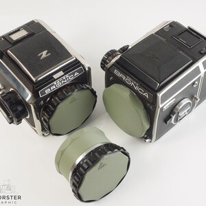 May include: Two black and silver Bronica Zenza Bronica cameras with green lens caps. A third green lens cap is in the foreground.