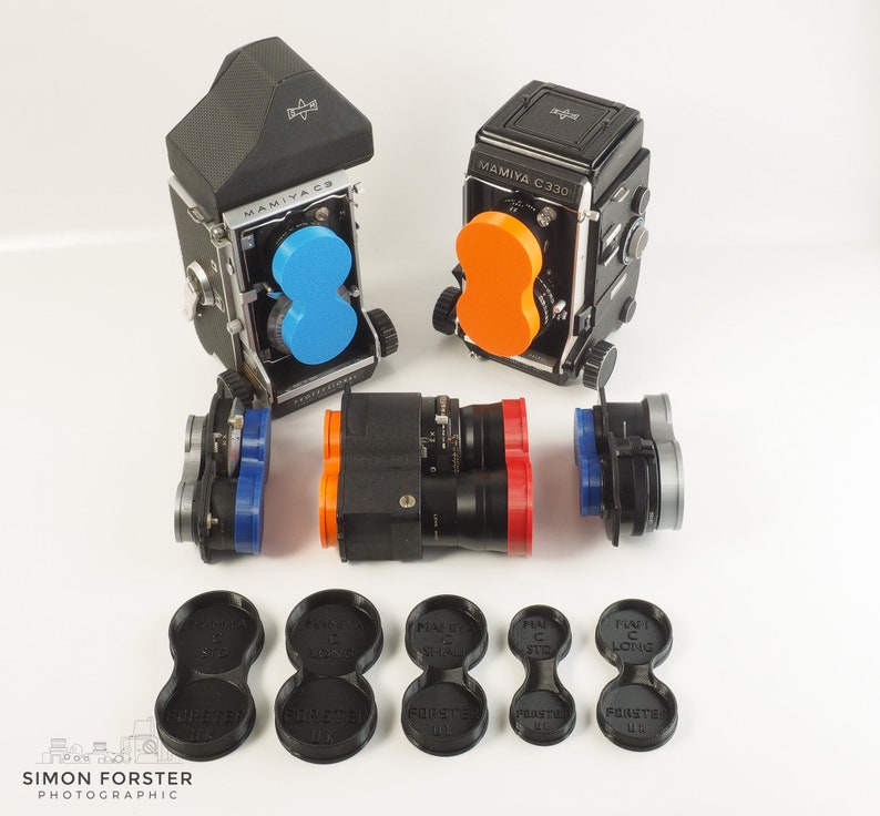 May include: Two vintage Mamiya C330 film cameras with interchangeable lenses, one with a blue filter and the other with an orange filter. The cameras are on a white surface with a variety of lens caps and filters.