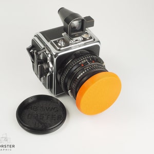 May include: A black and silver medium format camera with a bright orange lens cap. The camera has a red viewfinder and a black lens cap with the text "HB SWZ FORSTER LNY" printed on it.