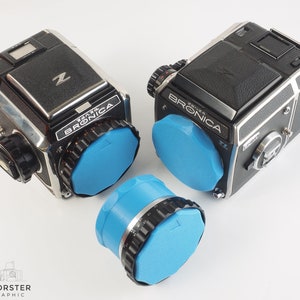 May include: Two black and silver Bronica ETRS cameras with blue lens caps. The cameras are on a white surface.