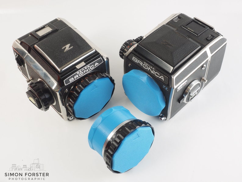 May include: Two black and silver Bronica ETRS cameras with blue lens caps. The cameras are on a white surface.