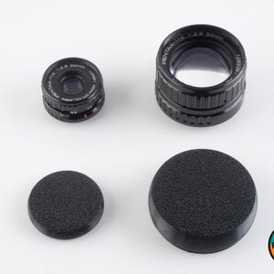 May include: Two black camera lens caps, one with a textured surface and one smooth, and two black camera lenses. The larger lens has the text "PENTAX-110 1:2.6 50mm 128936" printed on the side. The smaller lens has the text "PENTAX-110 1:2.8 24mm 123881" printed on the side.