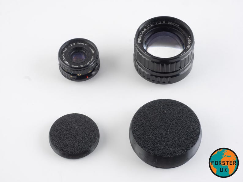 May include: Two black camera lens caps, one with a textured surface and one smooth, and two black camera lenses. The larger lens has the text "PENTAX-110 1:2.6 50mm 128936" printed on the side. The smaller lens has the text "PENTAX-110 1:2.8 24mm 123881" printed on the side.