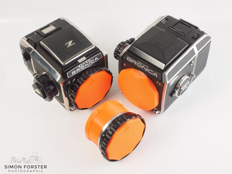 May include: Two black and silver Bronica cameras with orange lens caps. The cameras are on a white surface. The camera on the left has the text "Zenza Bronica" on it. The camera on the right has the text "Zenza Bronica ETR" on it.