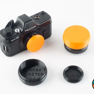 May include: A black Pentax auto 110 camera with a bright orange lens cap. Three additional lens caps are shown: one orange and two black. The black lens caps have the text "Pentax" and "Forster UK" printed on them.
