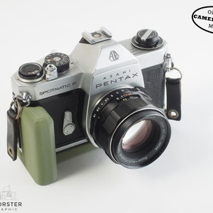 Peut inclure: Un appareil photo argentique 35 mm argenté et noir avec un objectif noir. L'appareil photo est doté d'une sangle en cuir noir et d'un étui d'appareil photo vert. L'appareil photo est marqué "Asahi Pentax Spotmatic F" et l'objectif est marqué "Super-Multi-Coated Super-Takumar 55mm f/1.8".