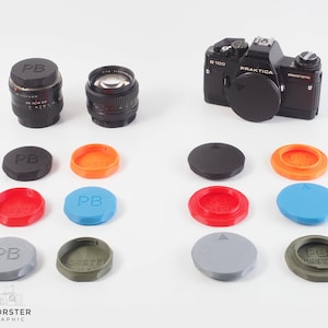 Könnte beinhalten: Eine Sammlung von Kameraobjektiven und Objektivdeckeln. Die Objektive sind schwarz mit silbernen Details. Die Deckel sind in verschiedenen Farben erhältlich: Schwarz, Orange, Rot, Blau, Grau und Olivgrün. Eine schwarze Praktica B100 Kamera.
