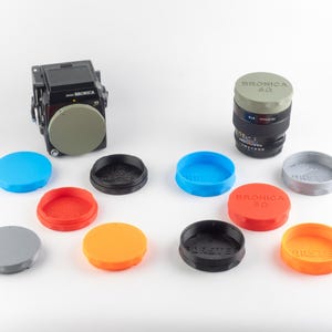 Op de afbeelding: Een verzameling camera-accessoires, waaronder een zwarte camera, een lens en verschillende gekleurde lensdoppen. De doppen zijn in verschillende kleuren: blauw, rood, oranje, grijs en zwart. De lensdoppen hebben de tekst "BRONICA SQ" erop.