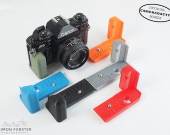 May include: A Canon AE-1 camera with a lens attached, surrounded by five 3D printed camera corner protectors in different colors: blue, black, gray, orange, and red. Each protector has the word "BUTTER" printed on it. The text "OFFICIAL CAMERADACTYL MAKER" is printed in a circle on the top right corner of the image.