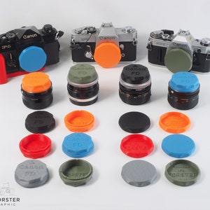 May include: A collection of camera lens caps in various colors, including blue, orange, green, and gray. The caps are labeled "Canon FD" and "FORSTER".