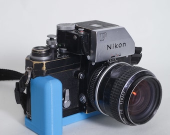 May include: A black Nikon F camera with a blue 3D printed camera grip attached. The camera has a silver lens with the text "Nikon" and "F" on the top. The lens has the text "1:2 928237" on the side.