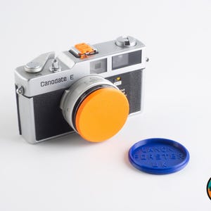May include: A silver Canonodate E camera with an orange lens cap. The camera is sitting on a white surface. A blue lens cap with the text "Canon Forster UK" is in the foreground.