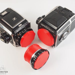 May include: Two black and silver Bronica ETRS cameras with red lens caps. The cameras are on a white surface.