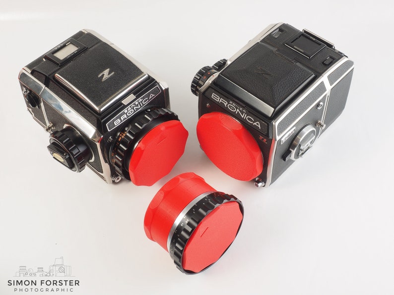 May include: Two black and silver Bronica ETRS cameras with red lens caps. The cameras are on a white surface.