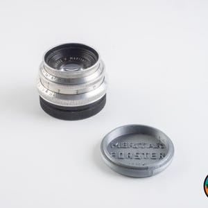 Op de afbeelding: Een zilveren camera lens met een zwarte lensdop en een grijze lensdop met de tekst "MEITAR FORSTER" erop gedrukt.