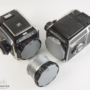May include: Two black and silver Bronica ETRS cameras with gray lens caps. One camera is on its side and the other is upright. A gray lens cap is in the foreground.