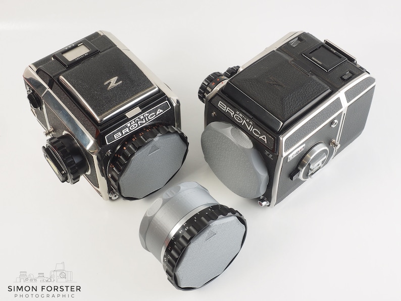 May include: Two black and silver Bronica ETRS cameras with gray lens caps. One camera is on its side and the other is upright. A gray lens cap is in the foreground.