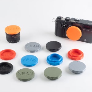 May include: A collection of 10 camera lens caps in various colours, including orange, grey, black, blue, and green. The caps are labelled "XPAN" and are arranged on a white surface. A black camera with an orange lens cap is also visible in the background.
