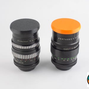 May include: Two black camera lenses with silver and white markings. One lens has a black cap, the other has an orange cap. The lenses have text that reads "Orestor 2.8/135" and "Pentacon 2.8/135".