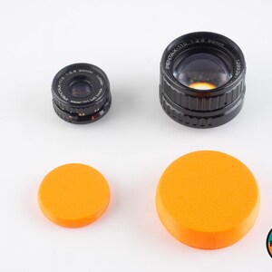 May include: Two black camera lenses with orange lens caps. The larger lens is labeled "PENTAX-110 1:2.4 50mm 1280357".