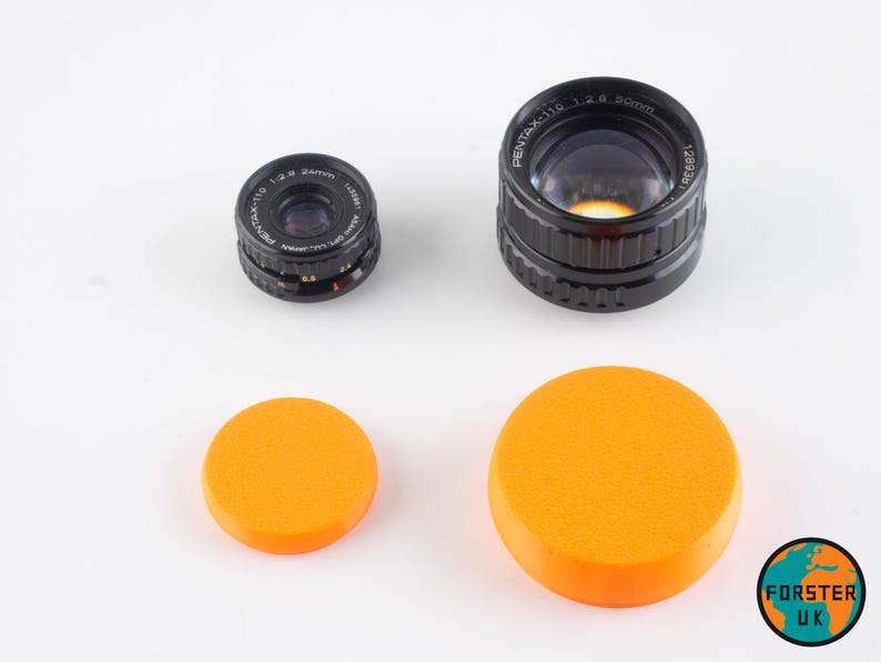 May include: Two black camera lenses with orange lens caps. The larger lens is labeled "PENTAX-110 1:2.4 50mm 1280357".