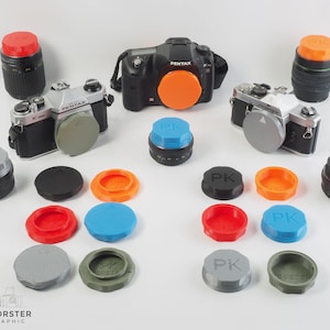 Op de afbeelding: Een verzameling van camera lensdoppen in verschillende kleuren, waaronder zwart, grijs, oranje, rood, blauw en groen. De doppen zijn gemarkeerd met de letter "PK" en zijn gerangschikt op een wit oppervlak. Er zijn ook twee camera's in de afbeelding, een zwarte en een zilveren.