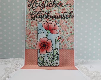 May include: A handmade greeting card with a floral design. The card features a pop-up element with red poppy flowers and green leaves. The background has a floral pattern and the words "Herzlichen Glückwunsch" in black script.