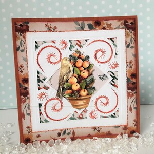 Puede incluir: Una tarjeta hecha a mano con fondo blanco y borde marrón. La tarjeta presenta un diseño recortado con un pájaro posado en un árbol en maceta con bayas rojas. El diseño está acentuado con costuras rojas.