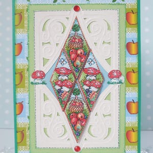 May include: A handmade greeting card with a green, white, and blue design. The card features a diamond-shaped design with a central image of a basket of fruit and a tree. The card is surrounded by a border of apples and leaves.