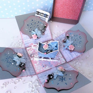 Könnte beinhalten: Eine blaue und rosafarbene Papierbastelbox mit einem Schneeflockenmuster. Die Box ist mit Blumen, Glitzer und einem kleinen blauen Schlitten verziert.