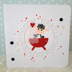 May include: A white card with a red and white circle with the text "GLÜCKWUNSCH HERZLICHEN" around the edge. Inside the circle is a black sheep holding a red heart in a red tub.