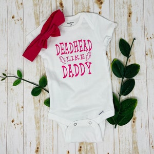 May include: White baby onesie with pink text that reads "Deadhead Like Daddy" with a lightning bolt graphic. A pink headband is included.