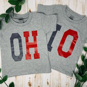 May include: Two gray t-shirts with the word "OHIO" printed in large, bold letters. The "O" is in dark gray, the "H" is in red, and the "I" is in dark gray.