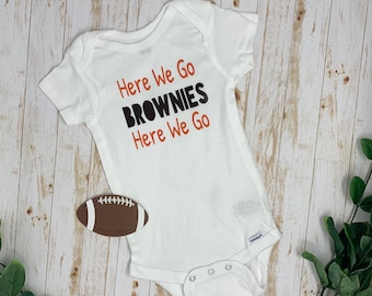 May include: A white short-sleeved baby bodysuit featuring the orange and black text Here We Go BROWNIES Here We Go printed on the front. A small brown and white football-shaped graphic is positioned to the left of the bodysuit.