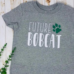 May include: A gray t-shirt with the words "FUTURE BOBCAT" in white block letters and a green paw print. The shirt is made of a soft material and is likely for a child.