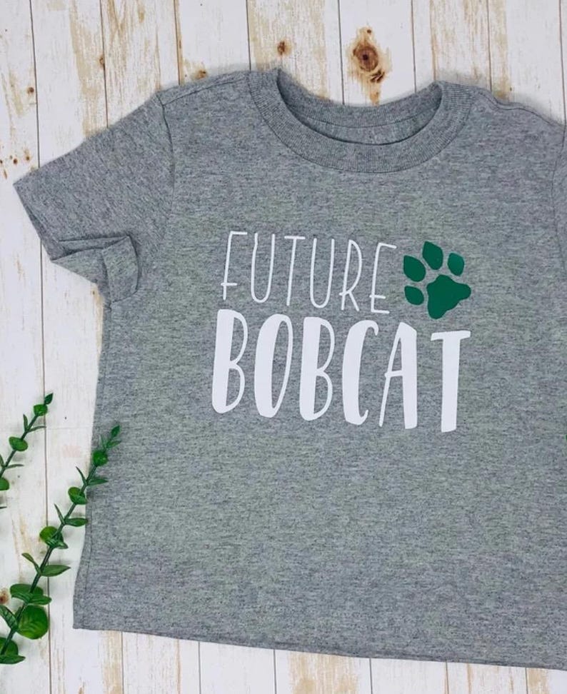 May include: A gray t-shirt with the words "FUTURE BOBCAT" in white block letters and a green paw print. The shirt is made of a soft material and is likely for a child.