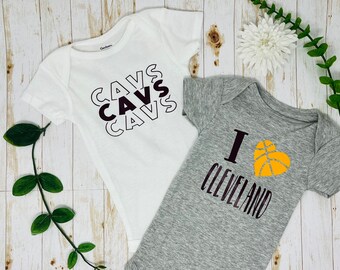 May include: Two baby onesies. One is white with the word "CAVS" repeated in a black and white design. The other is gray with the words "I CLEVELAND" and a basketball in a heart shape.