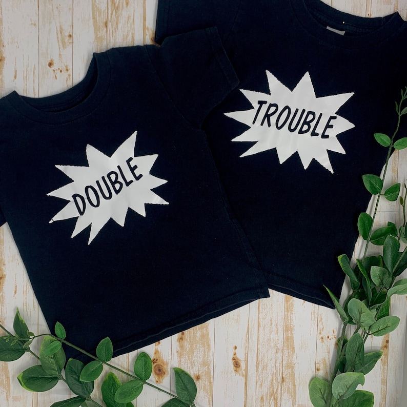 May include: Two black t-shirts with white text. One shirt says "Double Trouble" and the other says "Trouble".