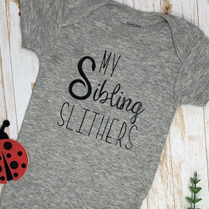 May include: Gray baby onesie with the text "My Sibling Slithers" in black. The onesie is made by Gerber.