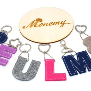 Könnte beinhalten: Eine Sammlung von Filz-Buchstaben-Schlüsselanhängern in verschiedenen Farben, darunter Marineblau, Lila, Grau und Pink, mit weißen Nähten. Eine Holzscheibe mit dem Wort "Monemy" ist ebenfalls vorhanden.