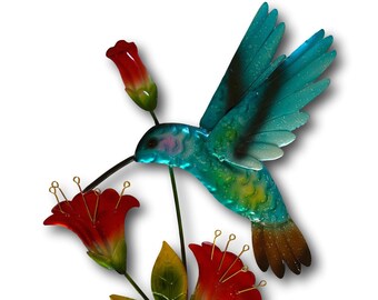May include: Metal wall art featuring a turquoise hummingbird with black markings, hovering near red flowers with green leaves. The hummingbird has a long beak and is depicted in flight.