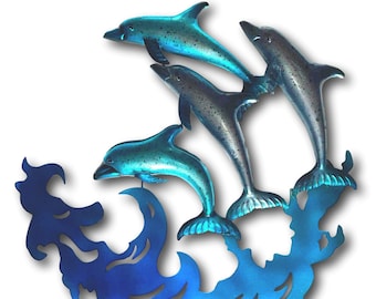May include: Metal wall art featuring four dolphins leaping above stylized blue waves. The dolphins have a metallic teal finish, and the waves are a darker blue. This decorative piece is designed to be mounted on a wall.