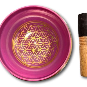 May include: A pink singing bowl with a gold flower of life design and a wooden striker.